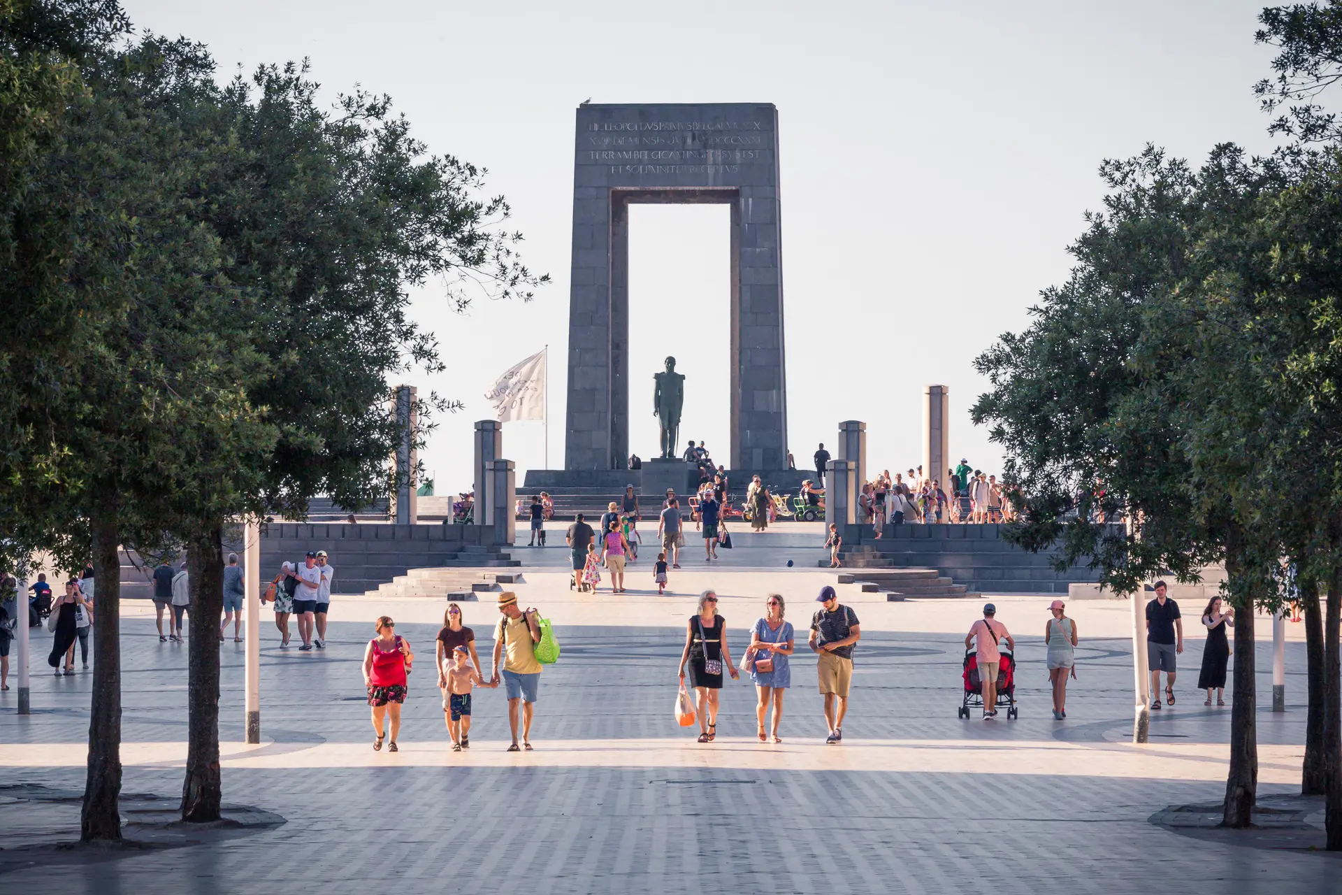 A bustling memorial site in De Panne, showcasing a monument and many visitors walking among trees on a sunny day.