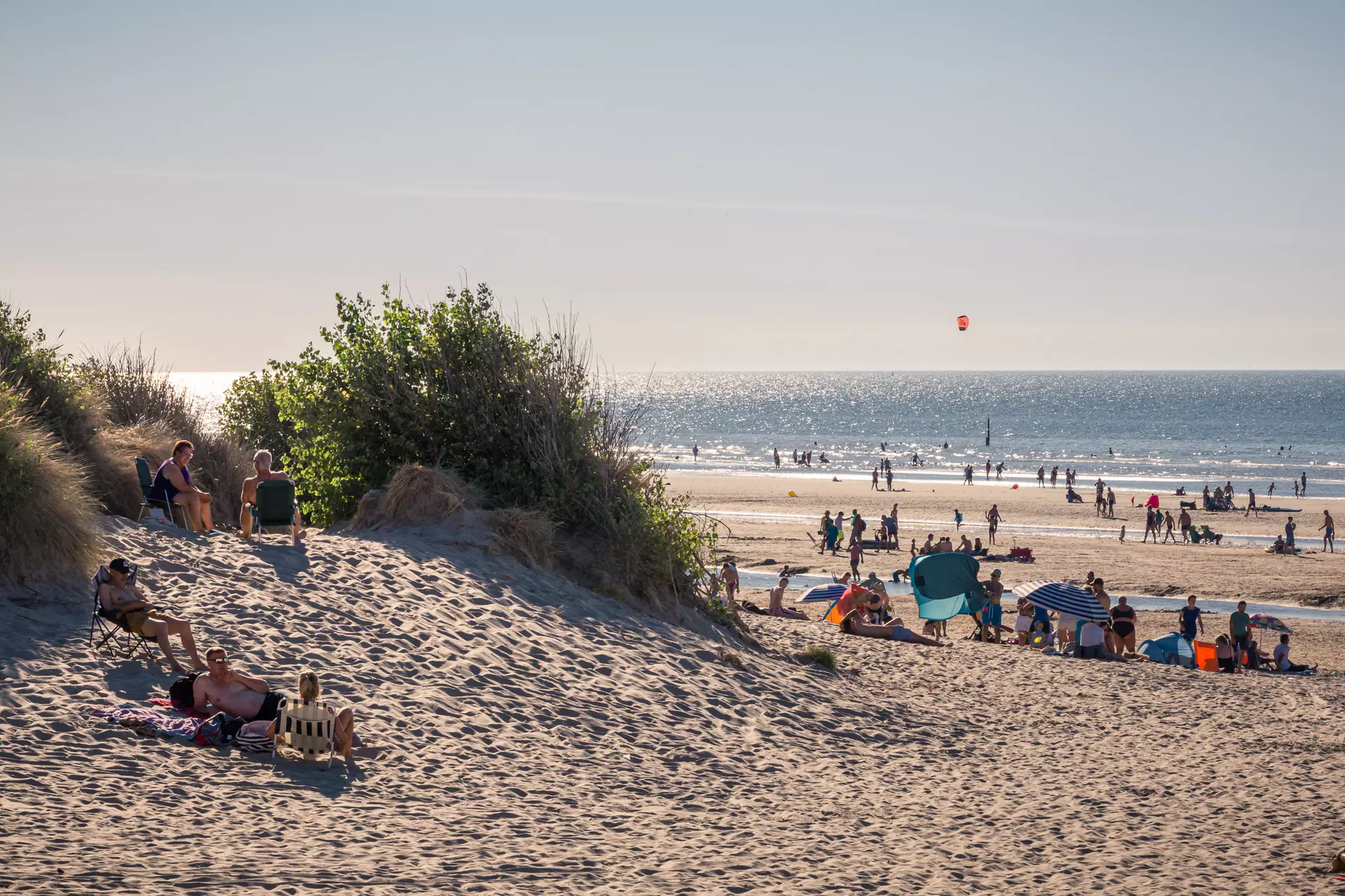 People relax on the sandy beach, some lying on blankets while others stand. The waves shimmer under the sun.