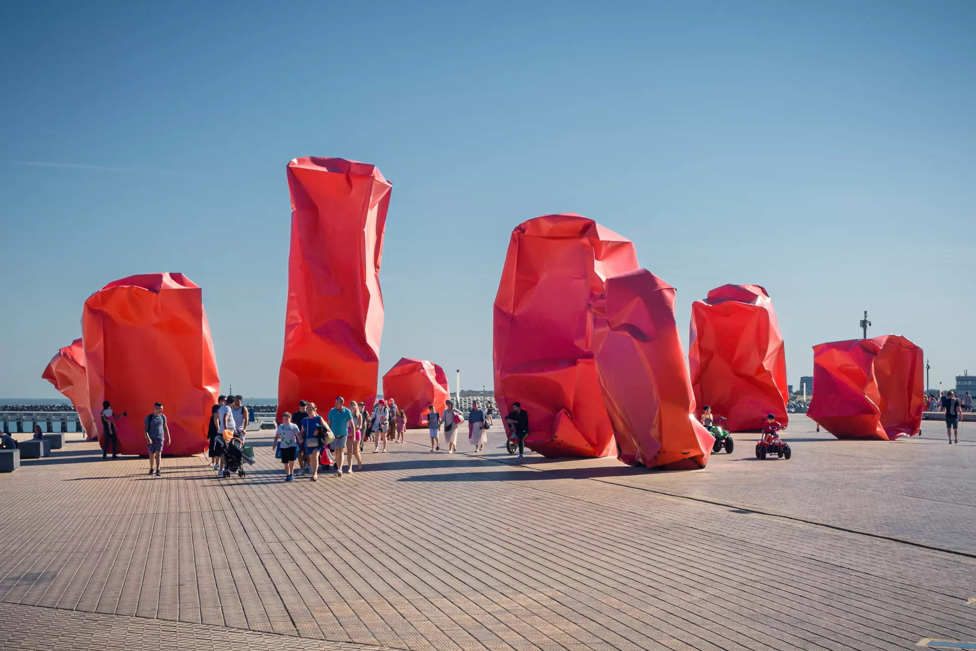 Large, red-orange sculptures stand in an open space as visitors walk around them in the sunlight.