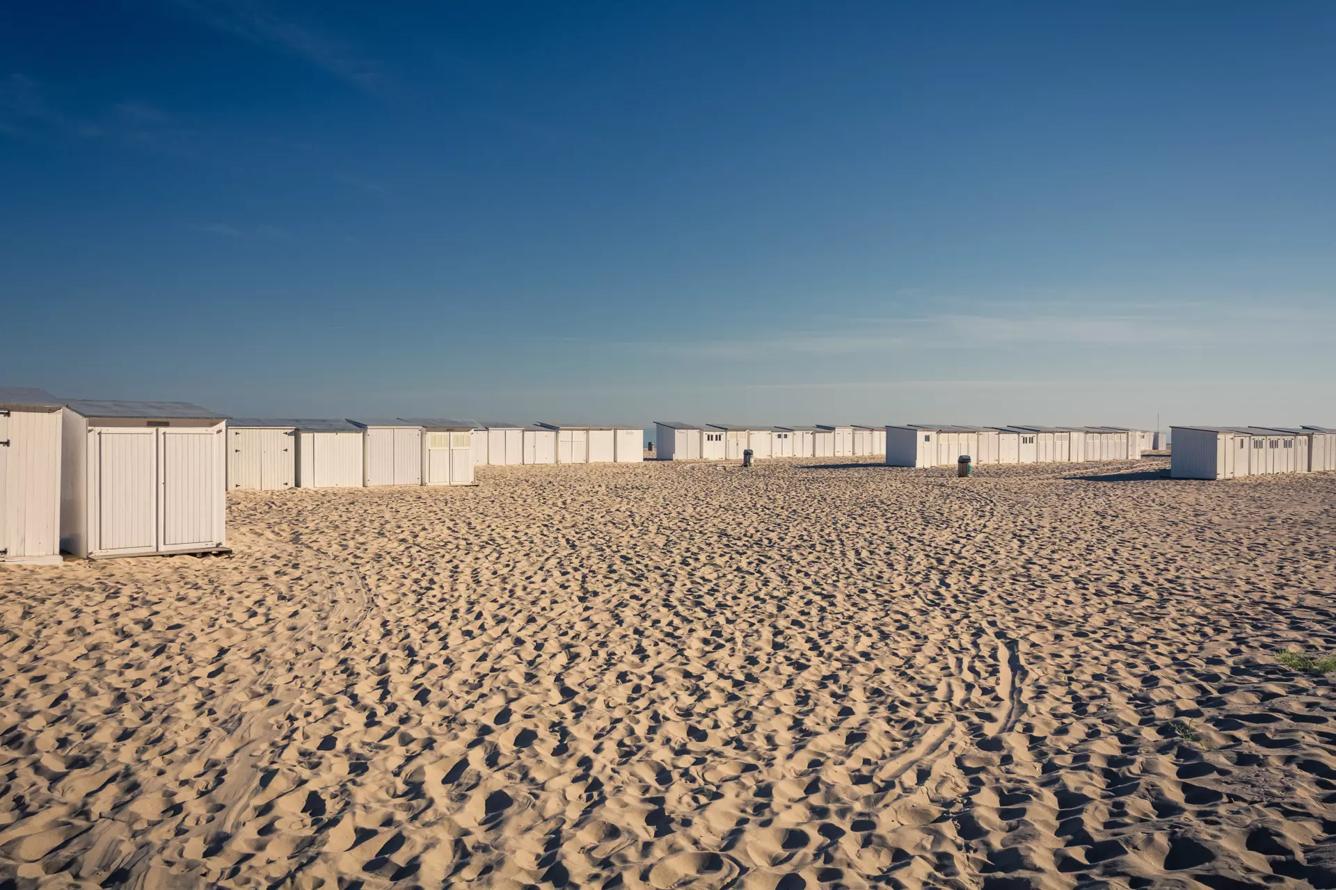 Bright sandy area with several white beach cabins arranged in a long line under a clear blue sky.