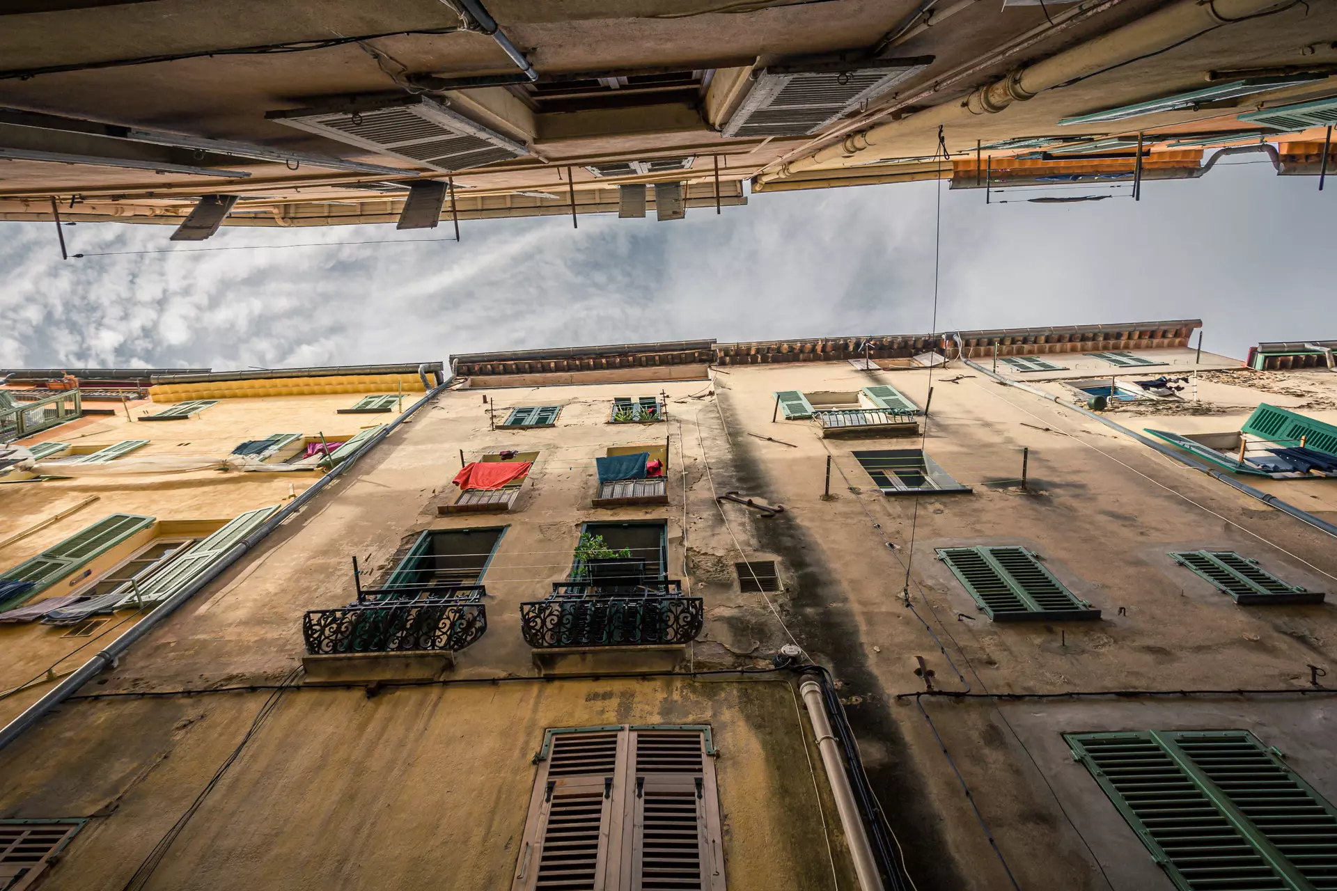 A long, narrow alleyway with old buildings, colorful windows, and laundry on balconies under a slightly cloudy sky.