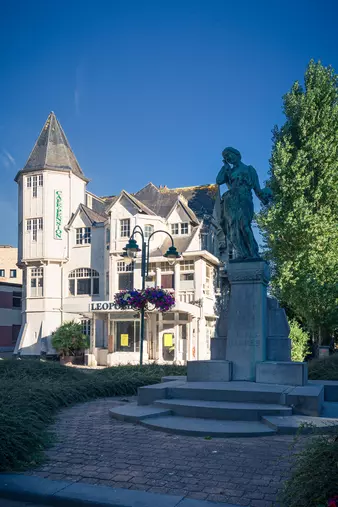 A statue of a mournful figure stands on a pedestal in front of a historic building with a tower and colorful flowers.