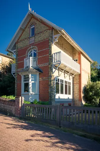 A bright, historic building with red and gray bricks, featuring a balcony and large windows surrounded by greenery.