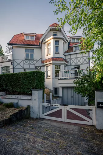 Ein mehrstöckiges Haus mit einem roten Ziegeldach und weißer Fassade, umgeben von Bäumen und einem Gartenzaun.