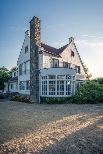 Ein mehrstöckiges, helles Haus mit Steinsäule und großen Fenstern, umgeben von trockenem Gras und Büschen.