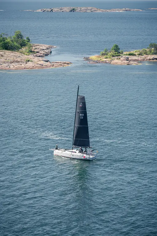 Segelboot mit schwarzem Segel fährt durch ruhiges Wasser, umgeben von kleinen, bewaldeten Inseln.
