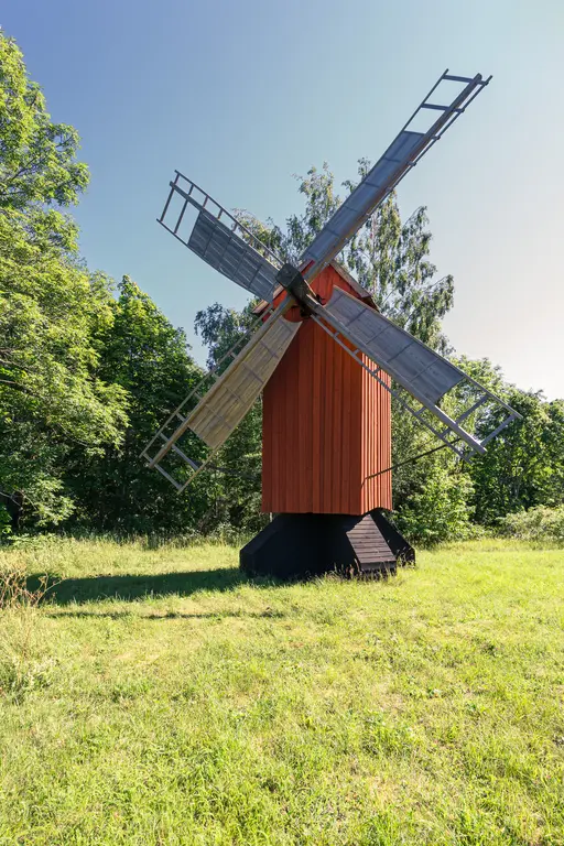 Rote Windmühle auf grünem Wiesenboden, umgeben von hohen Bäumen. Die Mühle hat ein quadratisches Fundament und große, sichtbare Flügel.
