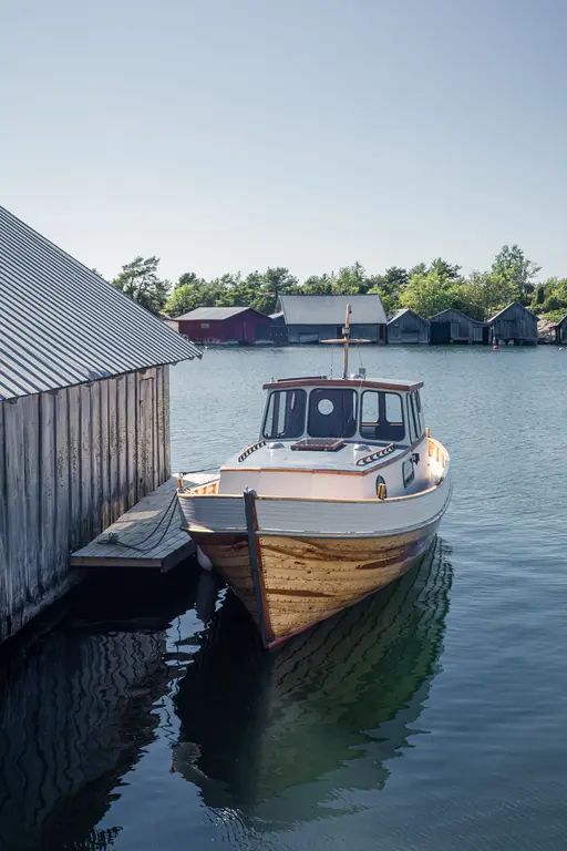 Boot am Anleger, umgeben von ruhig reflektierendem Wasser. Im Hintergrund stehen farbige Holzgebäude zwischen Bäumen. Klare Himmelsszene.