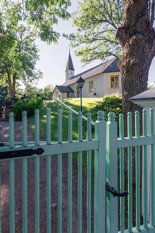 Eingang zu einem liebevoll gestalteten Garten mit grünem Zaun, Blick auf ein helles Holzhäuschen mit Kirchturm und umgebenden Bäumen.