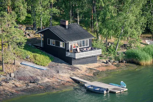 Ein schwarzes Holzhaus steht auf Felsen am Ufer, umgeben von Bäumen. Eine kleine Holzterrasse bietet Blick auf das Wasser, daneben liegt ein Boot.