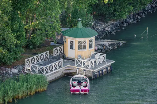 Kleine, rundliche Holzplattform am Wasser mit einem grünen Dach, umgeben von Bäumen und Steinen. Ein Boot liegt am Steg.
