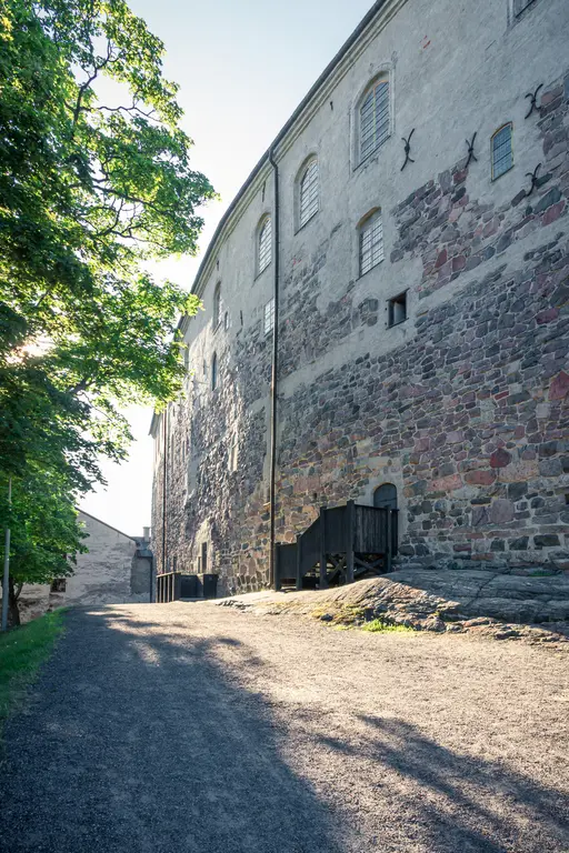 Mauer eines historischen Gebäudes mit einem Weg, der steil ansteigt. Im Vordergrund scheinen Schatten von Bäumen auf den Weg.