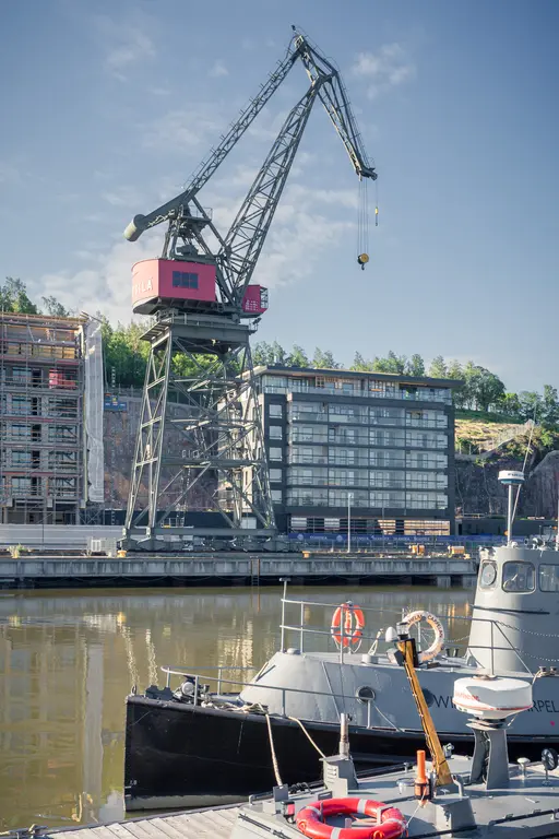 Hafenkran neben einem modernen Gebäude und einem schwarzen Boot im Wasser. Im Hintergrund sind Baustellen und Bäume zu sehen.
