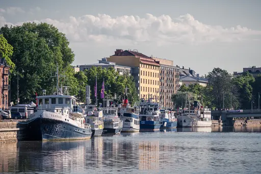 Boote liegen am pier, umgeben von grünen Bäumen und modernen Gebäuden. Ruhige Wasser spiegeln das Ufer wider.