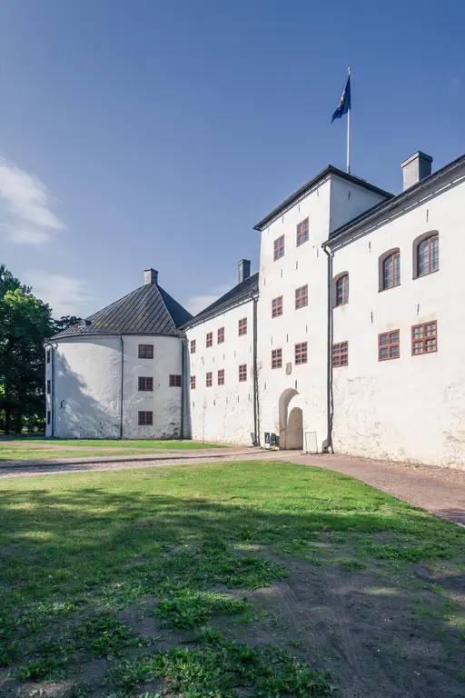Historisches Gebäude mit weißer Fassade, roten Fenstern und einer Fahne, umgeben von einem grünen Rasen und einem Kiesweg.