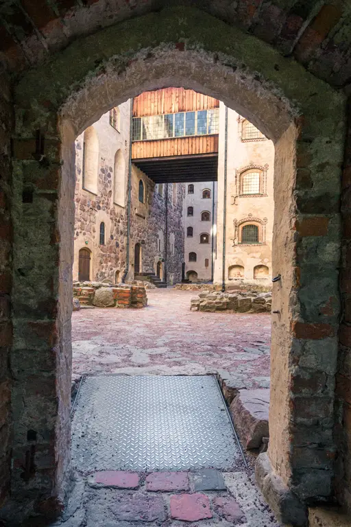 Durch einen alten Steinbogen sichtbare Innenhofszene mit historischen Gebäuden aus Steinen und Ziegeln sowie einem Holzsteg in der Mitte.
