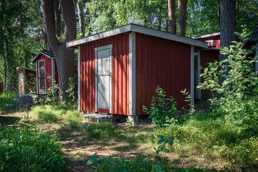 Rote Holzhütte im Wald, umgeben von Bäumen und Unterholz. Eine weitere Hütte ist im Hintergrund sichtbar.