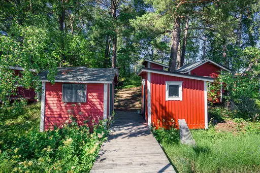 Rote Holzhütten zwischen üppigem Grün, umgeben von Bäumen. Ein Pfad aus Holzplanken führt hindurch.