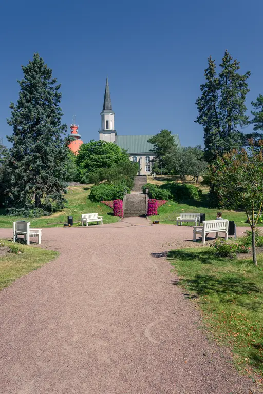 Parkansicht mit einem Weg, der zu einer Kirche mit spitzem Turm führt. Umgeben von Bäumen und Bänken, blühende Pflanzen zieren die Treppe.