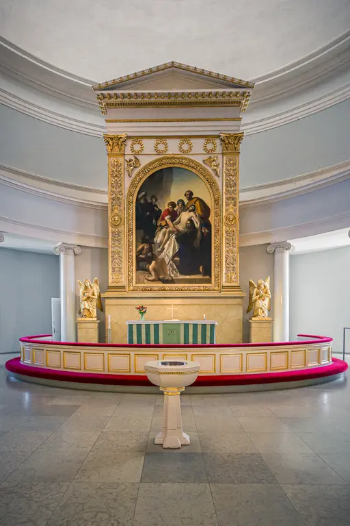 A church altar featuring a large painting, flanked by angel statues and a baptismal font, with a circular layout and decorative details.