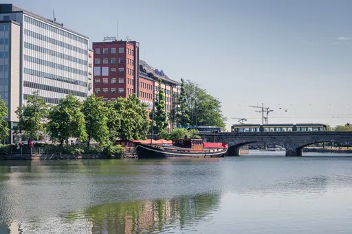 A scenic view of city buildings along a calm river, featuring a vintage boat and a bridge with a tram crossing. Lush greenery lines the waterfront.