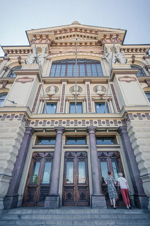 Historic building entrance with ornate wooden doors and classical architecture; two visitors approaching the steps.