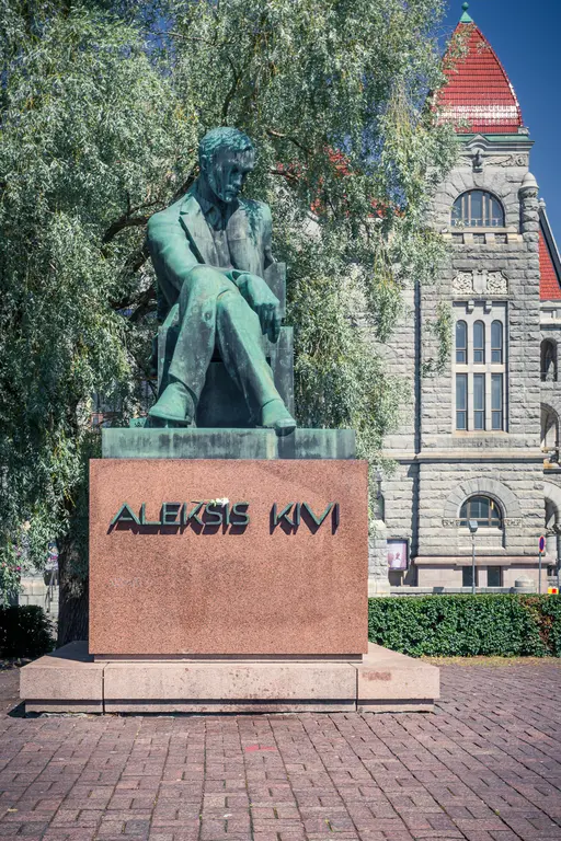 A bronze statue of a seated man deep in thought, titled "Aleksis Kivi," surrounded by greenery in an urban setting.