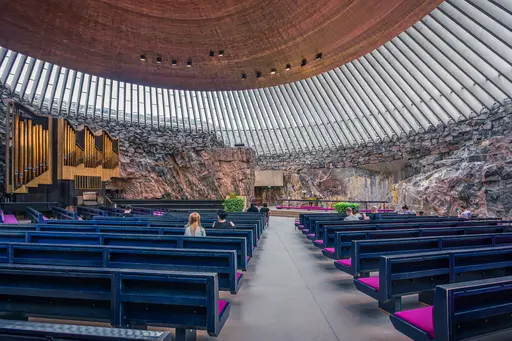 Interior of a modern chapel with a wooden, curved ceiling and rock walls. Pews arranged for worshippers, featuring greenery and organ pipes.