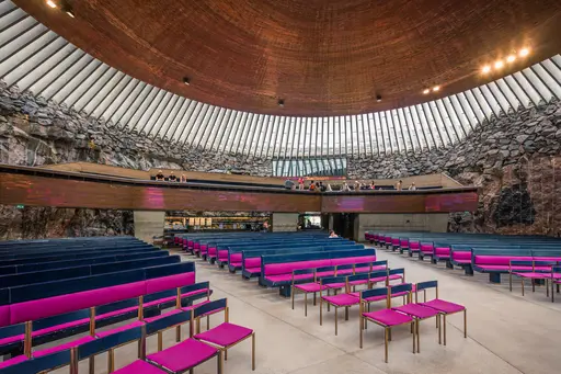 Interior of a modern rock church featuring rows of vibrant pink chairs, a wooden ceiling, and glass-walled natural light.