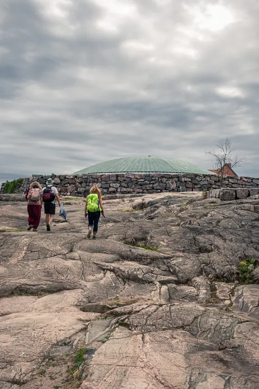 Hikers walking on rocky terrain leading to a unique stone structure under a cloudy sky in Helsinki.