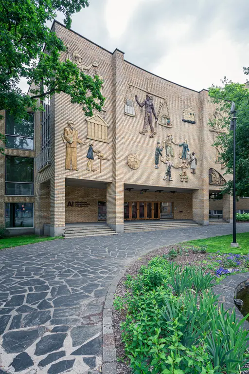 A distinctive brick building featuring artistic reliefs on its facade, surrounded by greenery and a stone pathway.