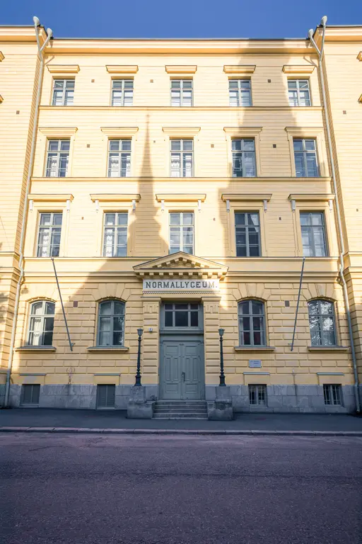 A historical yellow building with a green door and large windows, featuring a sign reading "NORMALLYCEUM." and lampposts.
