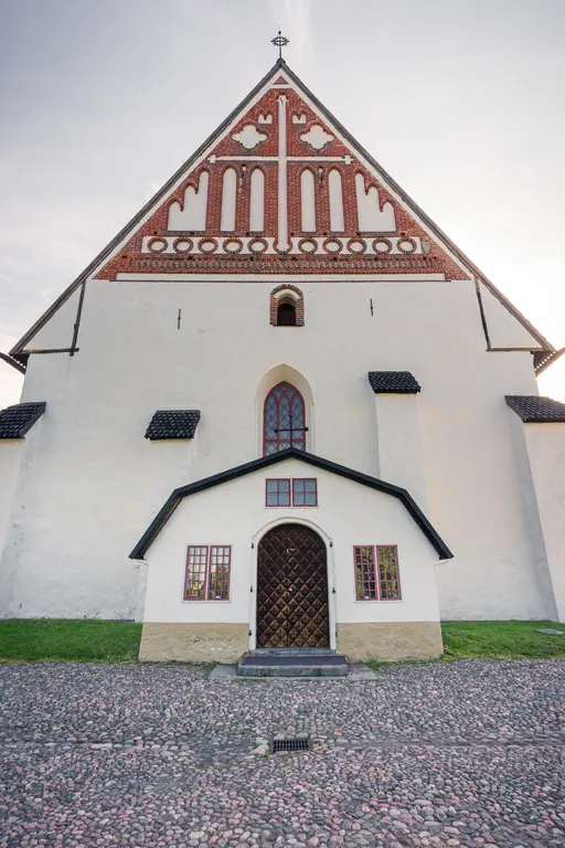 Fassade einer historischen Kirche mit einem weiß gestrichenen Unterbau und einem dreieckigen, gemusterten Dach. Eingangsbereich mit Holzporte.
