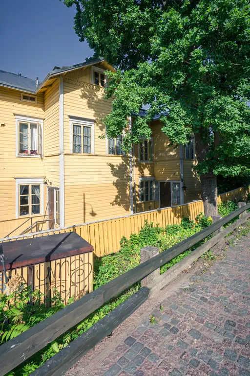 Gelbes Holzhaus mit vielen Fenstern und einem Kiesweg im Vordergrund, umgeben von Grünpflanzen und einem Holzzaun.