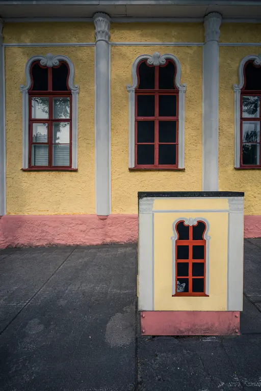 Fassade mit gelbem Putz und roten Fensterrahmen. An der Seite steht eine kleine Box mit einem Fenster, in dem ein Gesicht sichtbar ist.