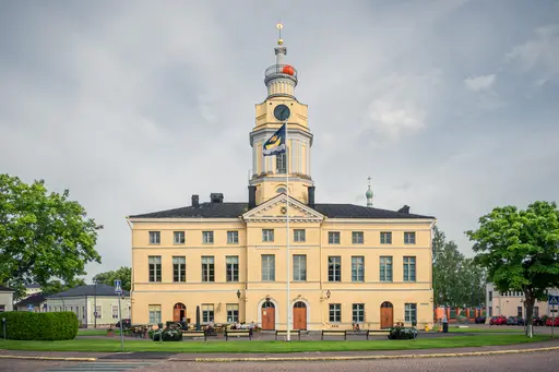 Historisches Gebäude mit gelber Fassade und Uhrturm, umgeben von Bäumen und einem kleinen Platz vor der Eingangstür.