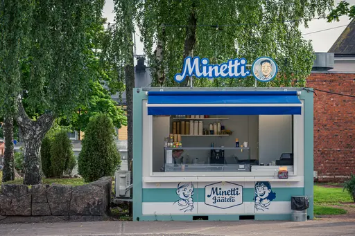 Eisverkaufsstand in Hamina mit blauem Dach, auf dem "Minetti" steht. Der Stand ist umgeben von Bäumen und hat eine Theke mit Eiscreme.