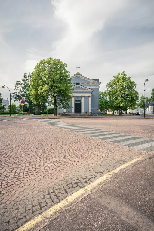Kirche mit Kreuz und Säulen, umgeben von Bäumen. Vor der Kirche verläuft ein gepflasterter Weg mit Zebrastreifen.