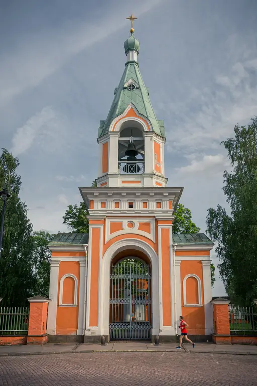 Historisches Tor mit grünem Dach und Glockenturm, umgeben von Bäumen; eine Person in rotem Shirt joggt vorbei.