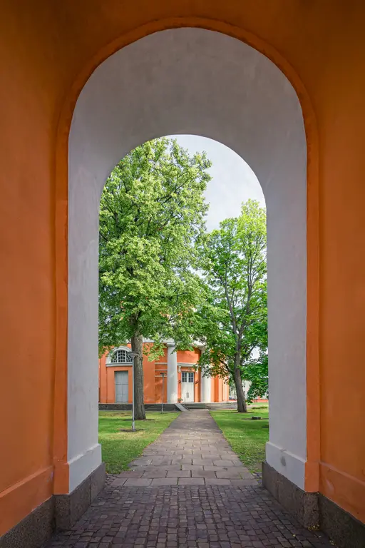 Durch eine bogenförmige Öffnung sieht man einen gepflasterten Weg, der zu einem orangefarbenen Gebäude führt, umgeben von grünen Bäumen.