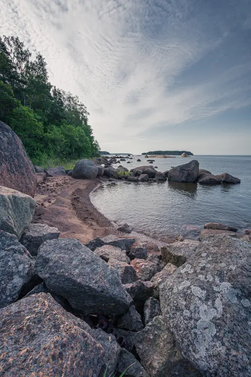 A rocky shoreline in Virolahti featuring smooth stones, gentle waves, and lush greenery along the coast under a cloudy sky.