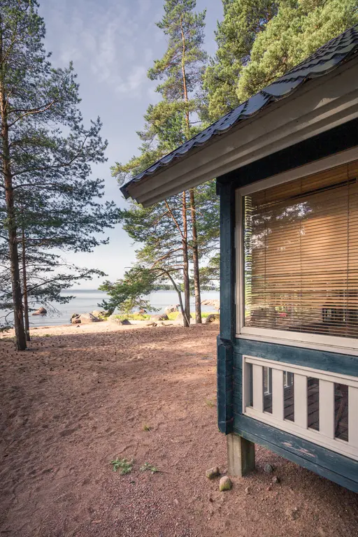 A cozy cabin in Virolahti, surrounded by sandy terrain and trees, with a view of the calm sea in the background.