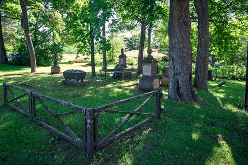 Historischer Friedhof mit Grabsteinen, umgeben von Bäumen und Grünflächen. Ein rustikales Zaunelement umrahmt den ruhigen Bereich.