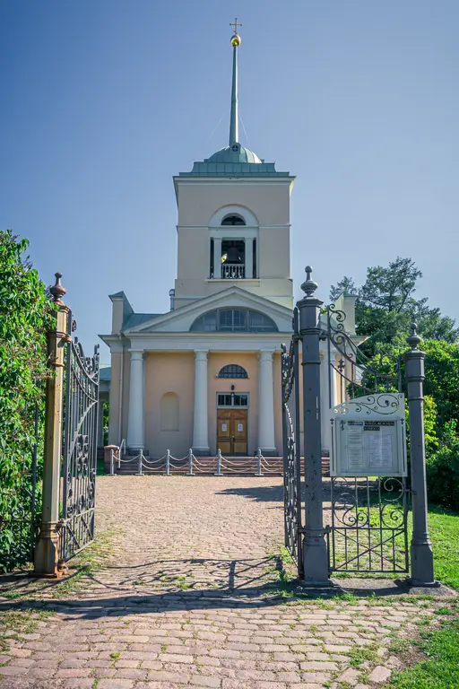 Eingang zu einer Kirche mit einer hohen Spitze und einem vergoldeten Kreuz. Der Weg ist von geschmiedeten Toren und grünen Bäumen umgeben.