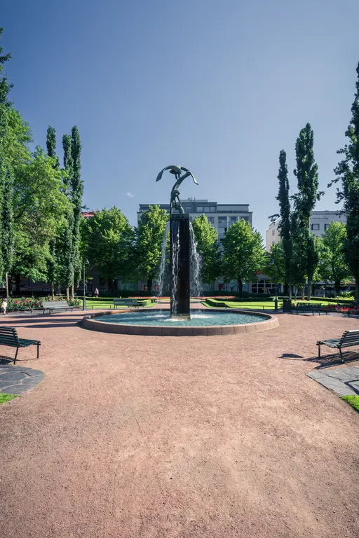 Brunnen mit einer Statue in der Mitte, umgeben von Bäumen und Blumenbeeten. Bänke stehen im lehmigen Bereich rund um den Brunnen.