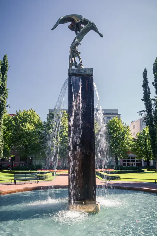 Brunnen mit Skulptur, das Wasser strömt über eine hohe, rechteckige Säule in einen runden, flachen Wasserspiegel. Grüne Bäume und Bänke umgeben die Szene.