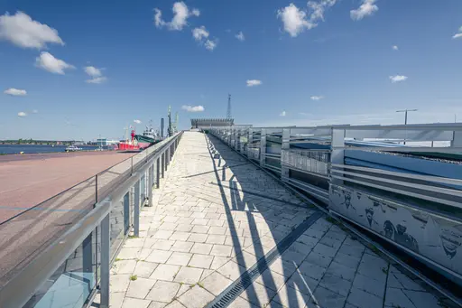 Blick auf einen gepflasterten Weg, der zu einem erhöhten Aussichtspunkt führt. Links sind Boote und der Hafen sichtbar, dazu ein klarer blauer Himmel mit wenigen Wolken.