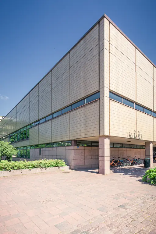 Moderne Gebäudearchitektur mit klaren Linien und einer gefliesten Fassade. Im Vordergrund stehen Fahrräder und Zierpflanzen.