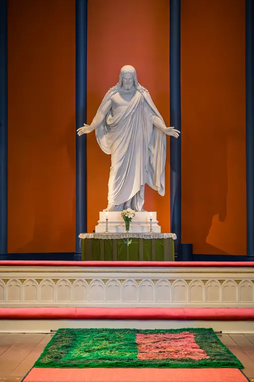 Skulptur von Jesus Christus in einem Kirchenraum, umgeben von orangefarbenen Wänden und einem altarähnlichen Tisch mit Dekoration. Vor dem Altar liegt ein buntes, gemustertes Teppichstück.