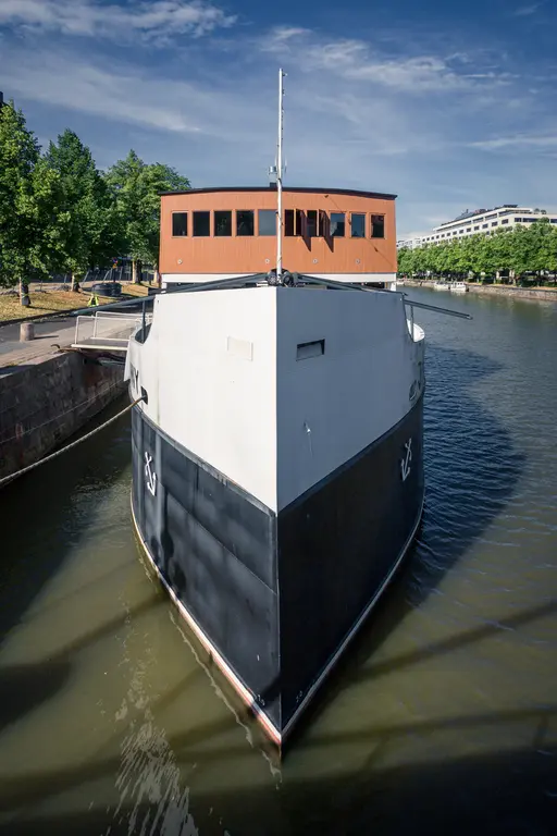 Blick auf die Vorderseite eines Schiffes mit einem hölzernen Oberdeck. Das Wasser spiegelt die Umgebung wider, während Bäume im Hintergrund sichtbar sind.